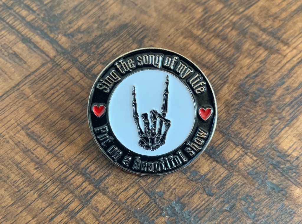 Custom round remembrance lapel pin for a funeral/celebration of life with a rock on skeleton hand that says "Sing the song of my life, put on a beautiful show" with two red hearts.